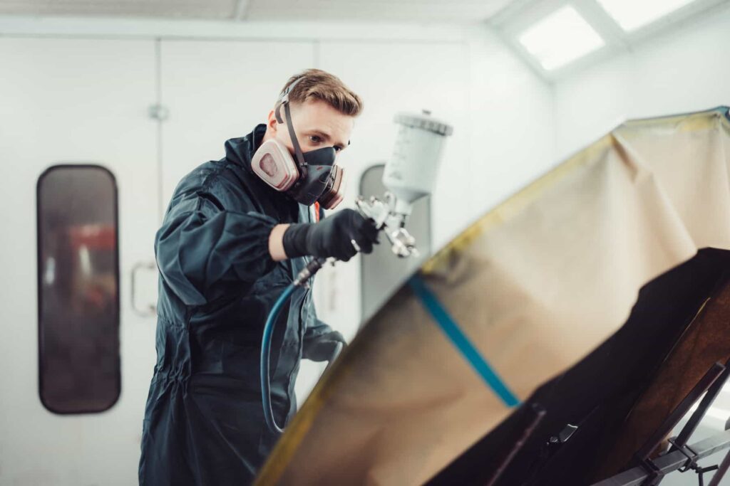 Ein Lackierer mit Atemschutzmaske und Overall hält ein Farbspritzgerät in einer Werkstatt und lackiert ein Fahrzeugteil.