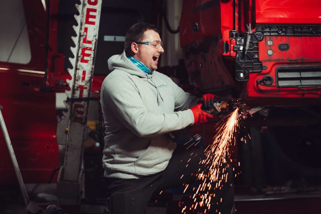 Ein Mechaniker hält eine Flex in einer Werkstatt und arbeit an der Karosserie von einem Auto. Es sprühen Funken.